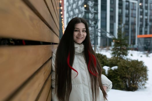 A woman is standing next to a wooden wall in a snow-covered environment. She is dressed warmly, with snowflakes falling around her. The scenery is serene and wintry, with a quiet, peaceful atmosphere.