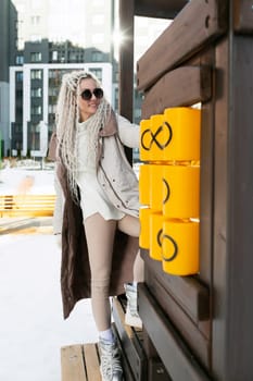 A woman stands leaning against a wooden wall, a coat draped over her shoulders. She appears contemplative, with a hint of relaxation in her posture.