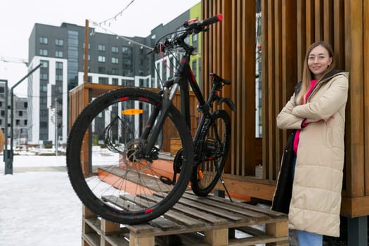 A woman is standing next to a parked bike on a wooden platform. She is looking towards the distance with her hand resting on the bike. The setting is outdoors with trees in the background.