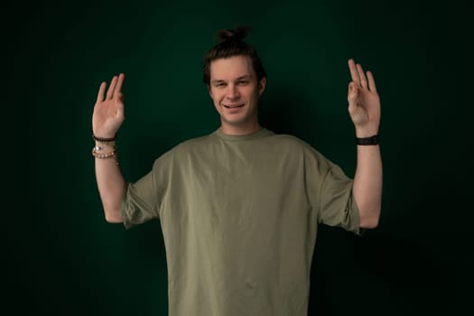 A man is standing in front of a solid green wall with his hands raised in the air. He appears to be in a pose of surrender or celebration.