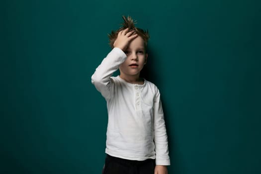A young boy is standing with his hands on his head against a vibrant green wall. He appears deep in thought or contemplation, with a simple and minimalist background.