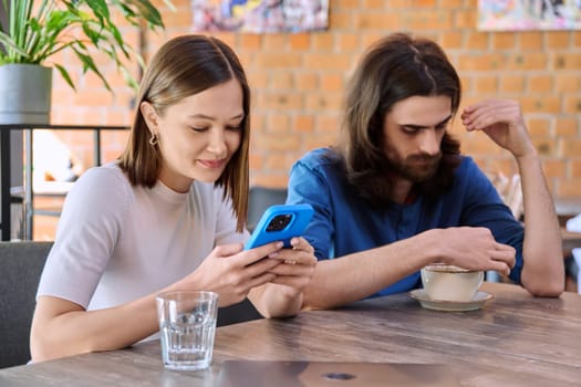 Young handsome people friends, couple man and woman relaxing together in a cafe, using smartphone, drinking coffee. Lifestyle, leisure, youth concept