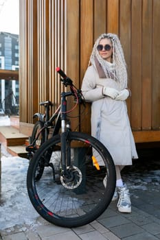 A woman wearing casual clothing is standing next to a bike parked on the sidewalk. The scene depicts a typical urban setting with the woman appearing to take a break from riding her bike.