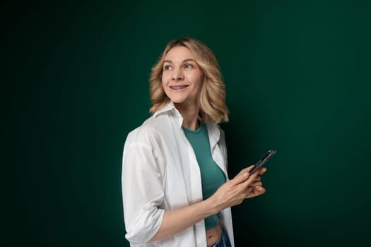 A woman is standing in front of a green wall, holding a cell phone in her hand.