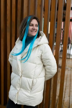 A woman with vibrant blue hair is standing confidently in front of a weathered wooden fence. She exudes a sense of individuality and strength in her pose.