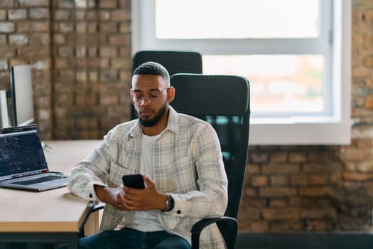 African American entrepreneur takes a break in a modern office, using a smartphone to browse social media, capturing a moment of digital connectivity and relaxation amidst his business endeavors