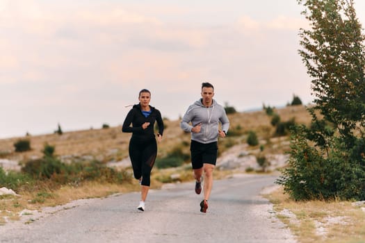 A couple dressed in sportswear runs along a scenic road during an early morning workout, enjoying the fresh air and maintaining a healthy lifestyle.