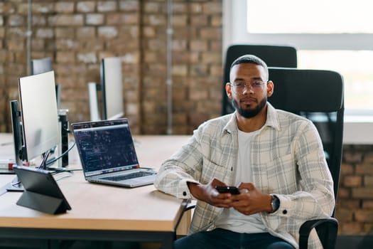 African American entrepreneur takes a break in a modern office, using a smartphone to browse social media, capturing a moment of digital connectivity and relaxation amidst his business endeavors