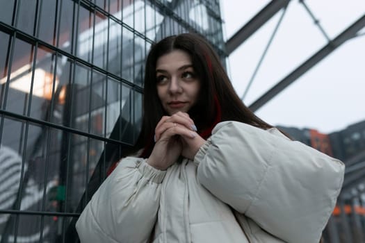 A woman is standing upright in front of a wooden fence, with her hands casually placed in her pockets. She appears relaxed and contemplative as she gazes ahead.