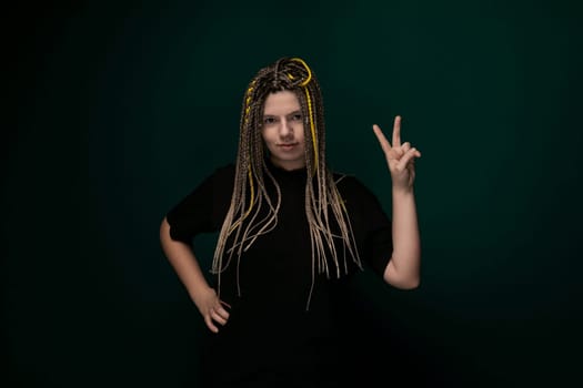 A woman with dreadlocks is extending her hand to make a peace sign gesture. She is dressed in casual clothing and is standing against a plain background.