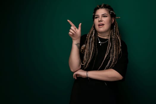 A woman with dreadlocks is standing in front of a green wall. She appears confident and stylish, with her hair cascading down her back. The background is a solid green color, creating a striking contrast with her dark hair.