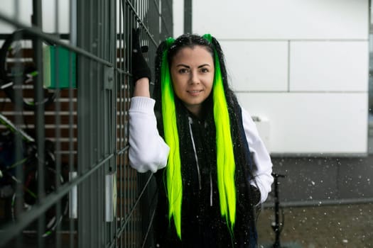 A woman with vibrant long green hair is standing next to a fence, looking off into the distance. She appears contemplative and thoughtful as she stands in the outdoor setting.