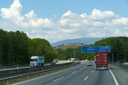 Hostalric, Spain - May 24, 2023: A large truck drives down a highway that runs parallel to a dense, verdant forest, providing a stark contrast between the man-made road and the untouched natural surroundings.