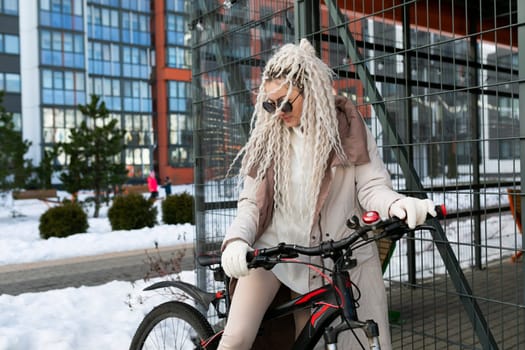 A woman with long white hair is riding a bike in the snow. She is bundled up in warm clothing as she pedals through the cold, snowy landscape.