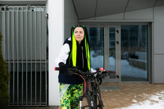 A woman is standing next to a bicycle in front of a building, looking towards the camera. The bike is parked on the sidewalk, and the background features the facade of a brick building.