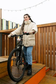 A woman is standing next to a bike on a wooden platform. She is wearing casual clothing and looking towards the distance. The bike is parked next to her, and the scene seems to be in a quiet outdoor area.