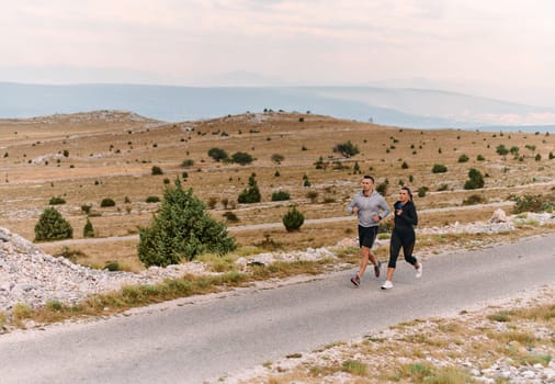 A couple dressed in sportswear runs along a scenic road during an early morning workout, enjoying the fresh air and maintaining a healthy lifestyle.