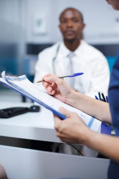 Close-up shot of a female patient filling out paperwork for her medical examination with the doctor at the hospital. Quality healthcare and advice are provided in this consultation.