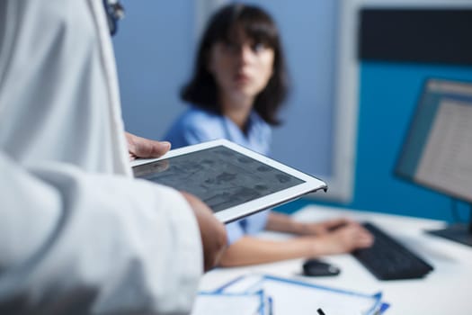 Selective focus on an individual wearing a white lab coat grasping a tablet while a nurse uses a desktop pc. Close-up shot of healthcare workers reviewing medical data by using modern technology.