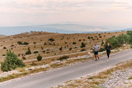 A couple dressed in sportswear runs along a scenic road during an early morning workout, enjoying the fresh air and maintaining a healthy lifestyle.