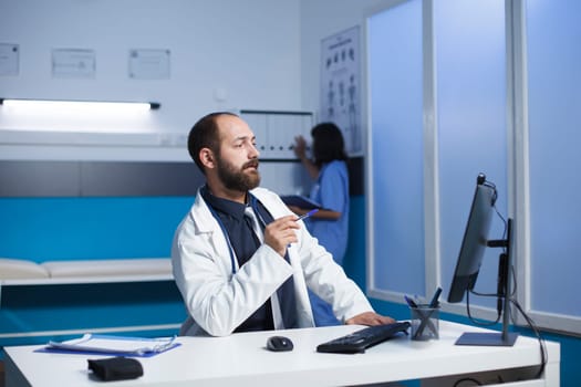 Doctor in a hospital communicates through a video call using his desktop computer. Caucasian man seated in the clinic office having a virtual meeting. Collaboration and technology in healthcare.