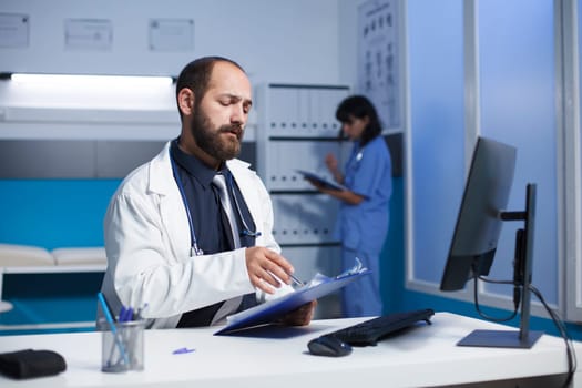 Image showcases a dedicated caucasian doctor checking and analyzing his notes at the clinic office desk. Young man wearing a white lab coat preparing for medical consultations with patients.