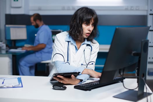 Female doctor checking patient records on computer while grasping a tablet. In an office, caucasian woman wearing a lab coat is using a digital device and desktop pc to analyze medical data.