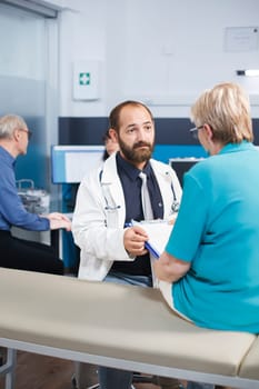 Elderly woman fills out medical forms and talks with doctor about her treatment. Portrait of male practitioner asking senior patient to sign documents for physiotherapy treatment.