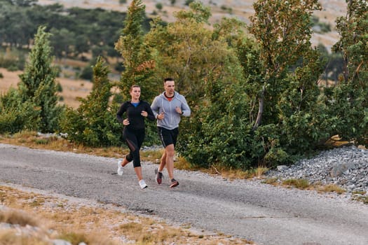 A couple dressed in sportswear runs along a scenic road during an early morning workout, enjoying the fresh air and maintaining a healthy lifestyle.