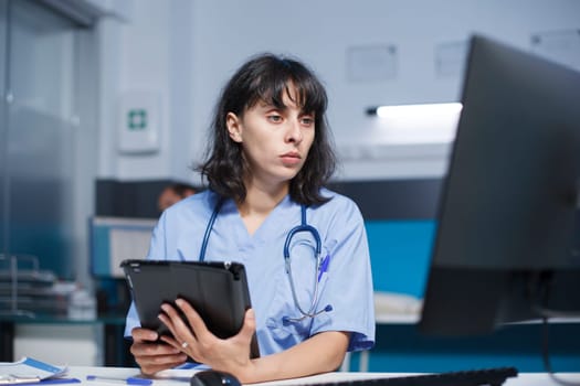 Caucasian nurse in medical uniform analyzing prescriptions for patients on tablet and computer, working in a hospital office. Physician assistance comparing healthcare data on digital devices.
