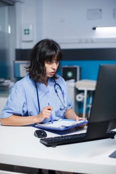 Focused female nurse is examining and rereading her notes at the clinic office room. When getting ready for patient medical consultations, caucasian practitioner uses desktop monitor and clipboard.
