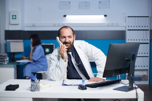 Caucasian male doctor seated at the office desk talking on the telephone with other healthcare specialists. Man wearing a lab coat is using a landline phone to speak with a hospital receptionist.