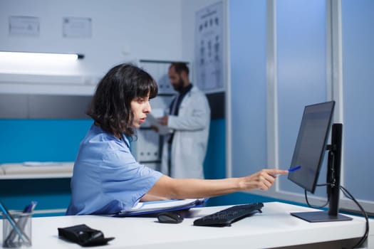 Image shows a nurse practitioner at the clinic desk reviewing and analyzing her notes. A Caucasian woman in blue scrubs prepares for patient medical consultations by using a desktop pc and clipboard.