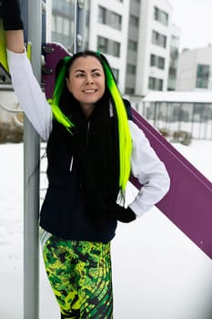 A woman with vibrant green hair stands next to a colorful purple and pink slide in a playground setting. She appears to be observing or contemplating the slides unique hues and design.