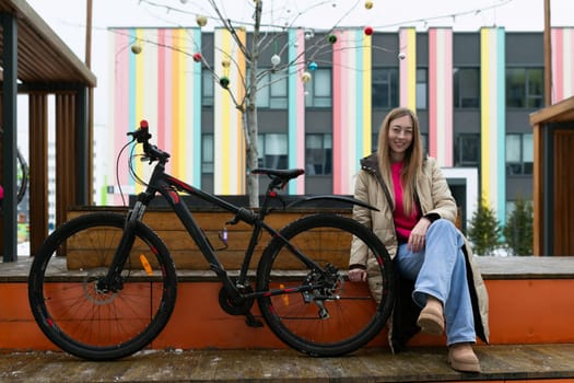 A woman in casual attire sits on a wooden bench placed beside a black bicycle. She appears relaxed and is gazing ahead. The bicycle leans against the bench, adding to the urban setting.