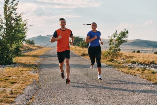 A couple dressed in sportswear runs along a scenic road during an early morning workout, enjoying the fresh air and maintaining a healthy lifestyle.