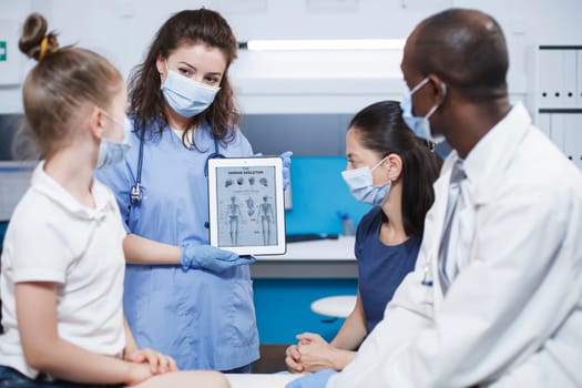 In clinic office, orthopedic doctor and nurse discuss the best treatment choices for the patient. Skeletal information is displayed on a tablet by a doctor wearing blue scrubs and a face mask.