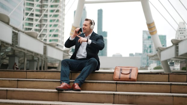 Professional business man listening headphone and using phone record voice while sitting at stair in urban city. Manager using headset listening relaxed song and moving along in lively mood. Urbane.