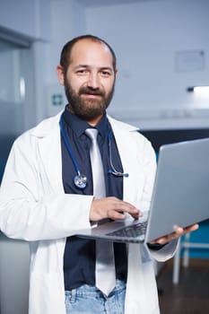 Portrait shot of a male physician in a white lab coat holding a digital laptop. Looking at camera is a caucasian doctor grasping a wireless computer in a hospital office room.