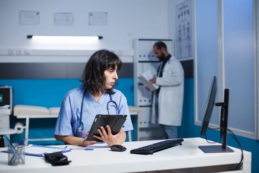 Female nurse uses a smart tablet and computer for medical information and checkups. While examining patient data on desktop pc, healthcare professional holds a gadget with a touch screen.