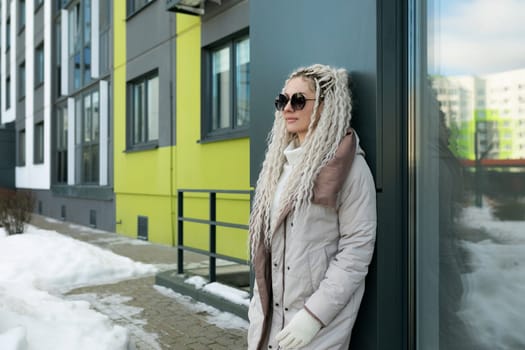 A woman with dreadlocks is leaning against a building. She appears relaxed, looking off into the distance. Her colorful clothing contrasts with the neutral tones of the building behind her.