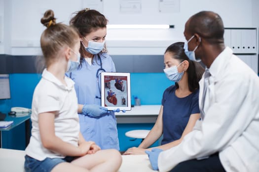 Black doctor shows cardiology image on digital tablet, pediatrician explains cardiovascular diagnosis to child and parent. Heart anatomy picture and heart disease problems presented by pediatrician.