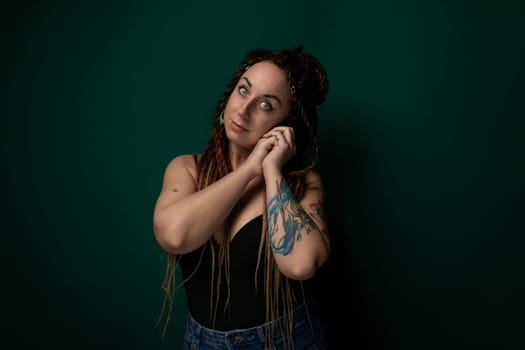 A woman with dreadlocks is casually leaning against a vibrant green wall. Her hair is styled in long, thin braids. She appears relaxed and confident, with a hint of contemplation in her expression.