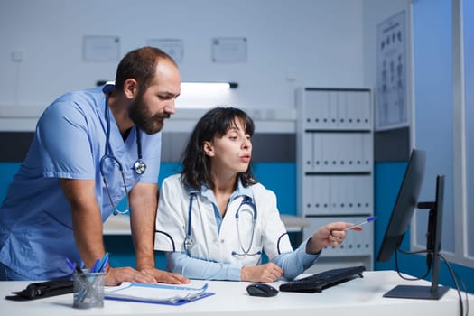 Caucasian medical staff in blue scrubs and lab coat collaborate digitally in a clinic office. They use a computer for conferencing, showing information and gesturing while technology dominates room.