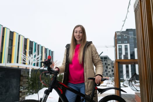 A woman with dark hair is standing beside a bicycle parked in the snow-covered ground. She is wearing a heavy winter coat and boots, looking towards the distance.