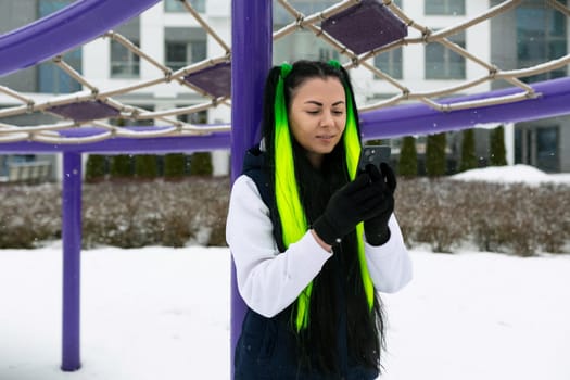 A woman with vibrant green and yellow hair stands confidently in front of a striking purple structure. Her bold hair colors contrast against the purple background, creating a visually arresting scene.