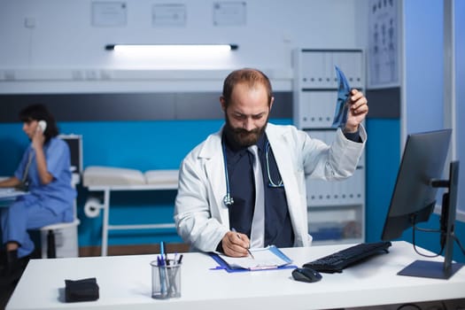 Dedicated medical doctor examines a CT image of a patient while taking notes on his clipboard. The image portrays a Caucasian male healthcare worker reviewing a chest X-ray imaging of a person.