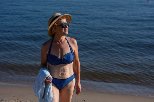 An old woman in a straw hat, sunglasses and a swimsuit is resting on the beach