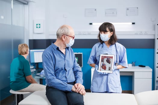 Senior man looking at heart illustration on tablet held by medical assistant. Female practitioner describing cardiovascular system and treatments to male elderly patient while wearing face masks.