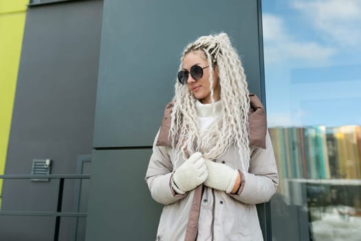 A woman with white dreadlocks is standing in front of a building. She is looking straight ahead, with a determined expression on her face. The building behind her is tall and features architectural details. The overall scene is urban and modern.